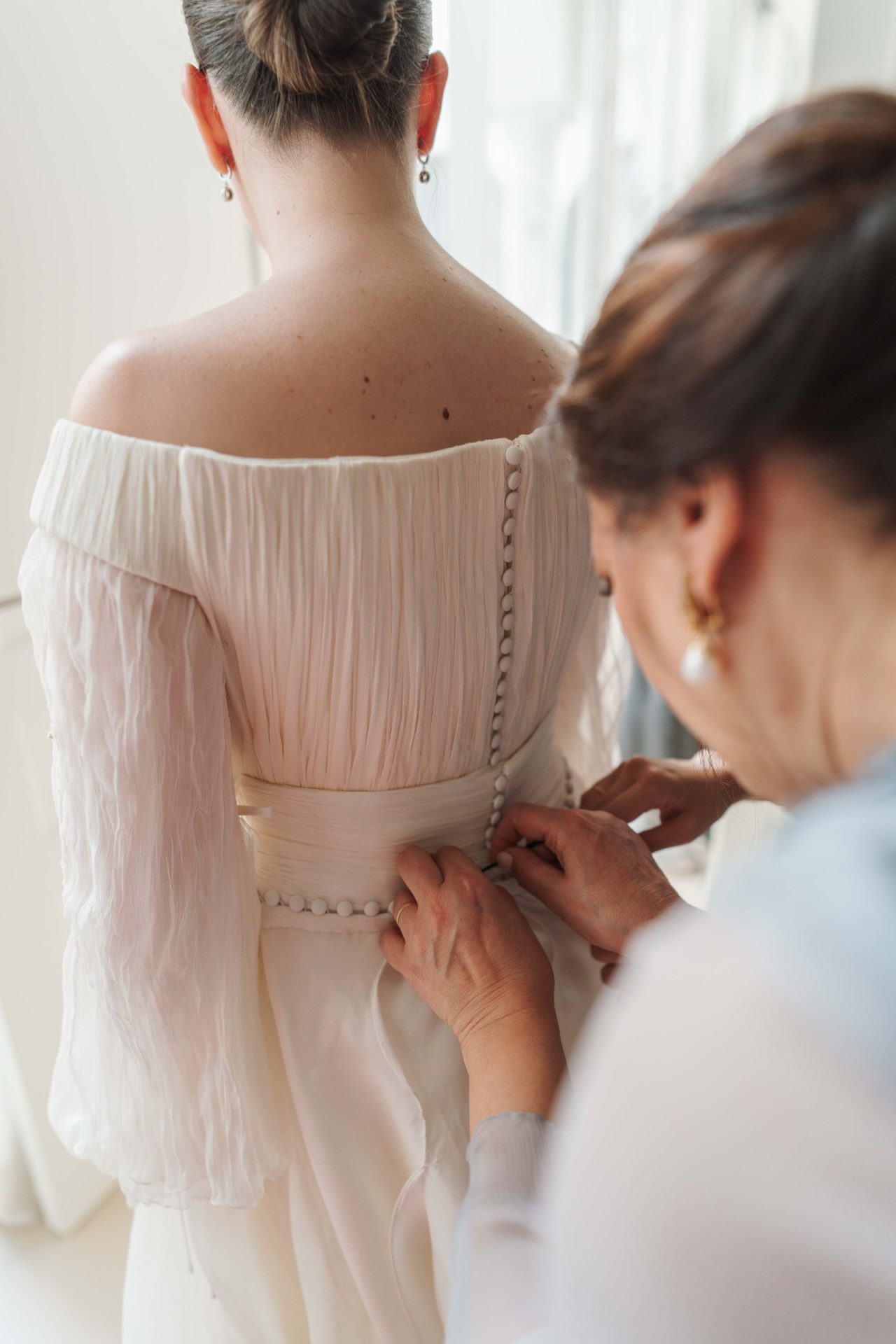 La madre de Inmaculada le abrocha los botones de la espalda; se aprecia el plisado de seda del cuerpo del vestido de novia, hecho a mano